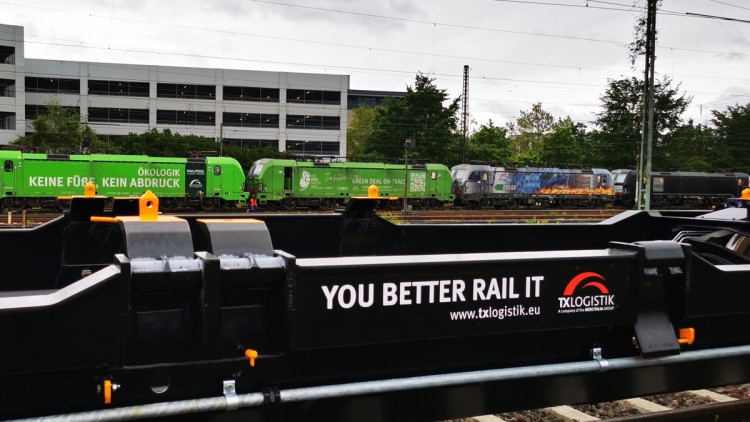 Ein leerer T3000-Waggon für Intermodaltransporte steht im Vordergrund. Auf den Gleisen im Hintergrund sind unter anderem grüne TX-Loks zu sehen
