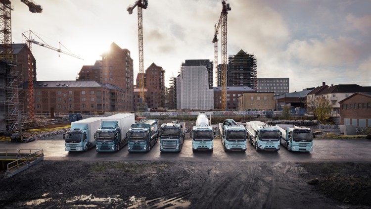 Acht türkisfarbene Elektro-Lkw von Volvo Trucks stehen aufgereiht vor einer Großbaustelle. Im Hintergrund sind mehrere Baukräne und mehrstöckige, zum Teil eingerüstete Gebäude in einer Großstadt zu erkennen